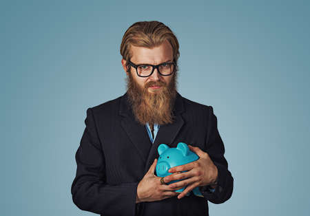 Young greedy stingy Bearded hipster businessman man holding piggy bank Isolated on blue studio wall Background. Negative face expression human emotion body language reaction attitude. Horizontalの写真素材