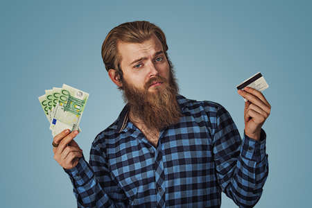 Businessman skeptical choosing holding showing plastic credit card or money cash euro banknotes bills Bearded hipster business man Isolated on blue Background. Negative face expression, human emotion.の写真素材