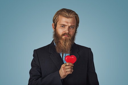 Serious Bearded hipster businessman man in black jacket and blue shirt holding red heart standing holding red heart shaped gift box and looking at camera Indoor studio shot isolated on blue backgroundの写真素材
