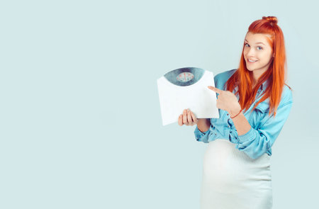 Content pregnant redhead girl in casual outfit holding weight scale and pointing at it smiling at camera isolated on light blue. Diet and weight care during pregnancy conceptの写真素材
