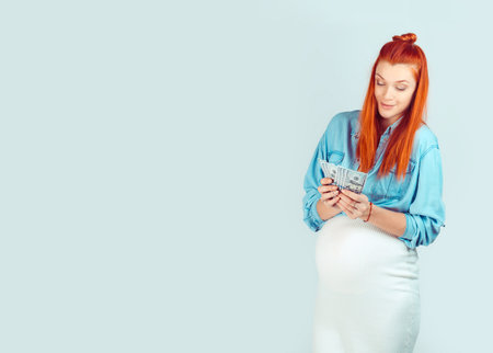 Content casual woman waiting for baby and holding pile of money for first time looking happy isolated on light blue. I received my start maternity benefit grant for pregnancy concept.の写真素材