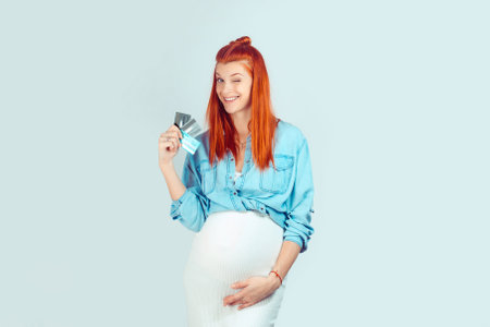 Beautiful ginger woman with belly holding few credit cards happy with money savings while smiling excitedly at camera on light blue backdrop. My cards are full even during pregnancy concept.の写真素材