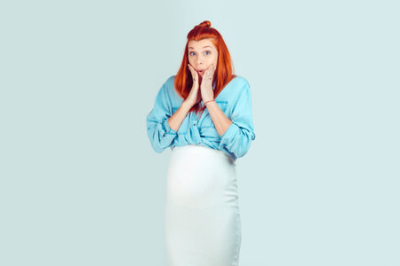 Happy adorable woman in expectancy embracing belly and touching chin looking away coquettishly while dreaming isolated on light blue. Omg, my water broke, I will soon have a babyの写真素材