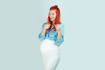 Happy redhead woman in expectancy pointing at camera with two fingers and winking happily isolated on light blue. I am pregnant, now is your turnの写真素材