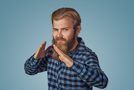 Closeup portrait of angry, mad, furious man raising hands in the air with karate chop. Hipster male with beard in blue plaid checkered shirt  Isolated blue studio Background. Negative face expression.の写真素材