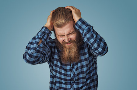 Depressed man having a very strong headache. Hipster male with beard in blue plaid checkered shirt  Isolated on blue studio Background. Negative face expression, human emotion, body language, reactionの写真素材