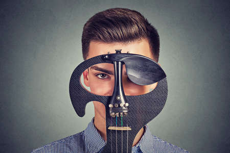 Young serious man with trendy hairstyle holding modern violin instrument looking through hole at cameraの写真素材