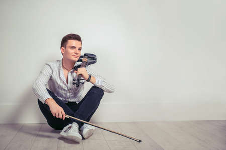 Handsome young man in shirt and denim holding violin and fiddle while sitting on white wall background and daydreamingの写真素材