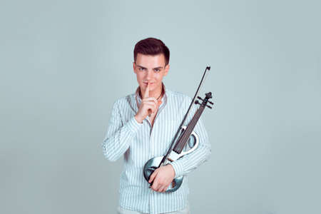 Young man in shirt holding modern violin and holding finger on lips asking for silence looking at cameraの写真素材