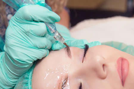 Micro pigmentation eyebrows work flow in a beauty salon. Close up woman having her eye brows tinted eyes closed. Semi-permanent makeup for eyebrows. Focus on model's face and tinted eyebrow right eye.の写真素材