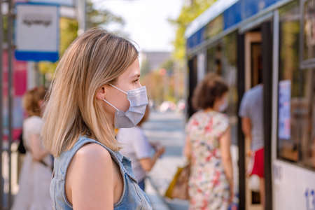 Cheerful woman wearing a sterile protective medical mask against coronavirus, Covid-2019 Asian pandemic sars virus at public bus station in European city street looking ahead, people on the backgroundの写真素材