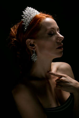 Beauty queen. Woman in profile with diamond crown on head touching nude shoulders with eyes closed isolated on dark grey background. Multicultural Caucasian Irish modelの写真素材