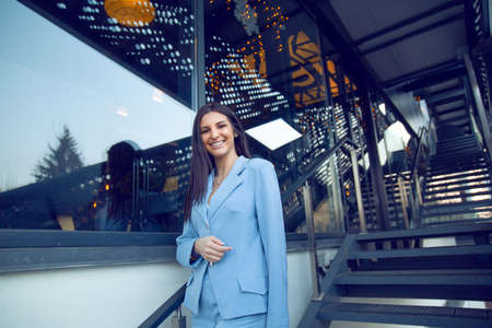 Cheerful woman wearing blue formal suit standing outside on metallic stairs on street urban...