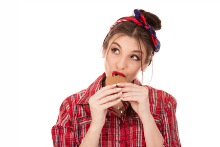 beautiful young woman eating bicuit cookies, in red checkered shirt, looking to side up isolated over pure white studio background, positive emotion, holding biscuits in hands, trying sweet foodの写真素材