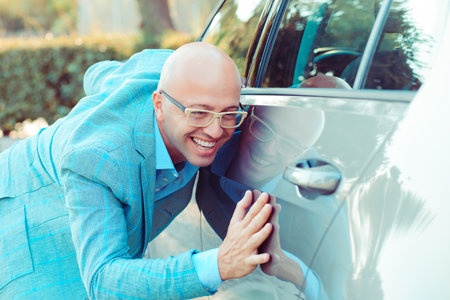 Happy excited young bald italian man driver embracing petting his car outdoors. Green energy biofuel electric environment friendly new car concept.  Positive face expression human emotion reactionの写真素材