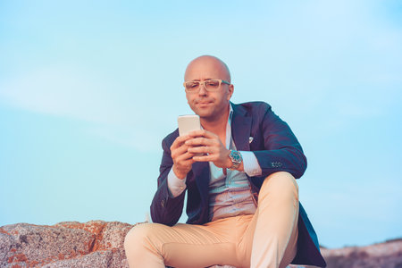 Closeup portrait puzzled confused business man looking at smartphone thinking reply to received sitting on rocks blue sky on background. Negative face expression human emotion reaction body languageの写真素材