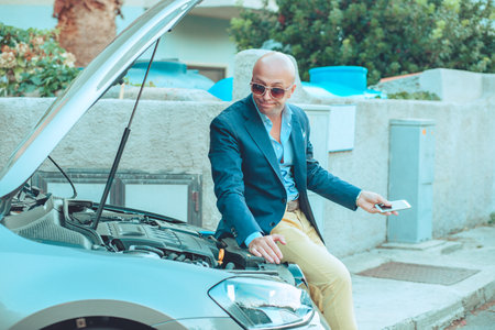 Young businessman man sitting on his car looking into open hood frustrated because of his broken car holding cell phone in hand outdoor urban road background. Positive face expression emotion reactionの写真素材