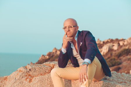 Handsome man outside thoughtful sitting on rocks outdoors blue sky on background. Negative face expression human emotion reaction body language attitudeの写真素材