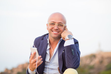 Happy guy business man in eyeglasses using a smart phone to listen music with headphones sitting outdoors blue sky on background looking at you Positive face expression emotion body languageの写真素材