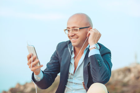 Handsome smiling businessman guy having fun listening to music on phone spending time outdoors sitting on rocks blue sky on background. Positive face expression human emotion reaction body languageの写真素材