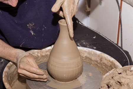 Pottery - skilled wet hands of potter shaping the clay of a vase on potter wheel. Pot, vase throwing. Manufacturing traditional handicraft bowl, jar, pot, jug.の写真素材