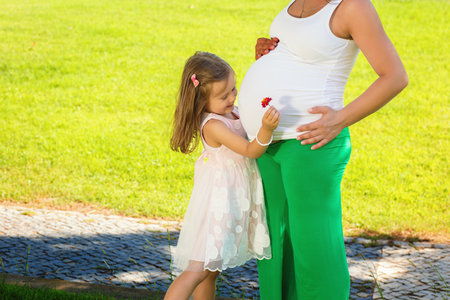 Cute little girl hugging and kissing her mothers pregnant belly in summer nature outdoors in a city parkの写真素材