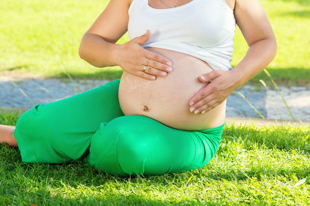 Young pregnant woman protecting her stomach with sunscreen cream before train while sitting in park on green grass meadow. Expecting woman applying cream.の写真素材