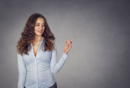 Closeup portrait happy confident young smiling woman gesturing, presenting pointing finger copy space at right isolated grey wall background. Positive human emotion sign symbol face expression feelingの写真素材