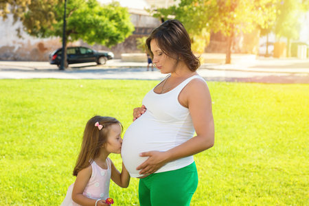 Cute little girl hugging and kissing her mothers pregnant belly in summer nature outdoors in a city parkの写真素材