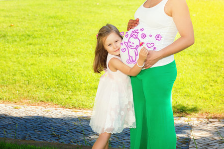 Cute little girl hugging and kissing her mothers pregnant belly in summer nature outdoors in a city parkの写真素材