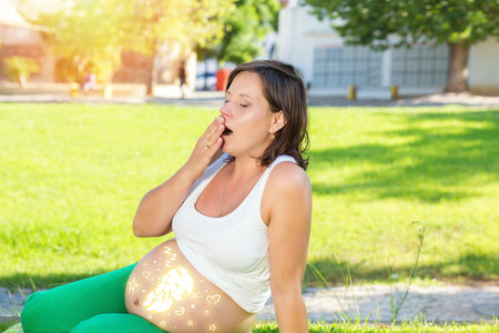 Pregnant woman yawning. Lovely Future Mother Prepares For Birth of Baby, Tired and Trying to Relax, sitting on a Green Grass in a Sunny Park Outdoors in Daytime. Girl With brunette bob Hair style.の写真素材