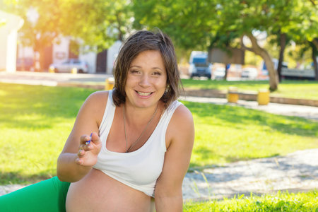 Smiling pregnant woman talking with distant client looking at camera, record shoot video training online training webinar video blog, sitting outdoors on green grass meadow in a city park on sunny dayの写真素材