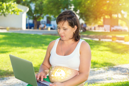 Happy pregnant woman working a laptop computer sitting outdoors on a green grass meadow in a city park on a sunny dayの写真素材