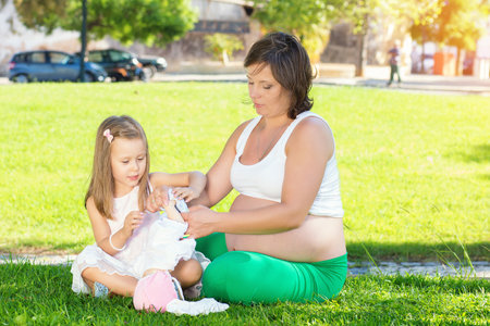 Kid girl withpregnant mom resting playing with a doll in park on green grass. Girls playing with toys. Both and wearing white clothing dresses. Picnic outdoors on warm summer day. Copy spaceの写真素材