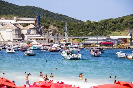 Beach on the coast of Brazil in Arraial do Cabo, the sand is white, the sea is turquoise and is crowded. It has red umbrellas and tables and chairs of the same color. People are sitting or going to get into the sea for swimming or playing around.のeditorial素材