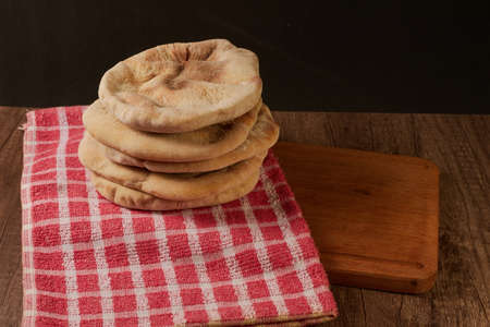several pita breads stacked on a rustic cloth and a cutting board on a wooden table with black...