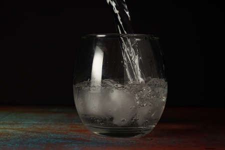 Glass of ice water being filled with sparkling water on a dark background and rustic surface.の写真素材
