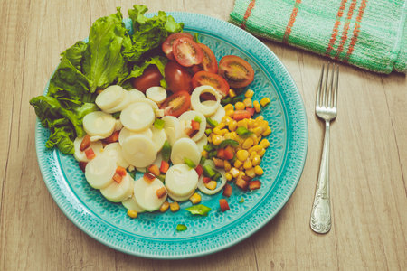 Fresh and exquisite salad of palm hearts cut into slices, cherry tomatoes, corn kernels, red pepper, lettuce in a vintage light blue plate on a wooden table ready to taste.の写真素材