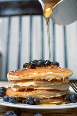 Stack of homemade pancakes prepared with blueberries and a jar with honey falling on the pancakes an...