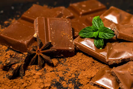 Different pieces of dark chocolate and cocoa powder on a dark surface. Mint leaves and star anise seeds complete the picture.の写真素材