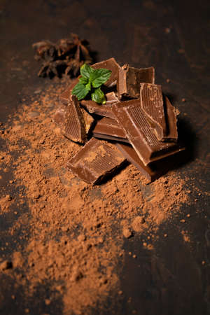 Different pieces of dark chocolate and cocoa powder on a dark surface. Mint leaves and star anise...