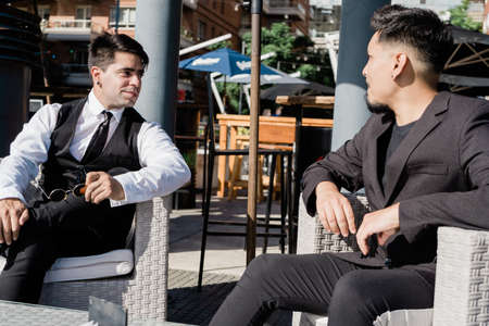 Young Latin American men in elegant clothes sitting in armchairs outside talking casually after office.の写真素材