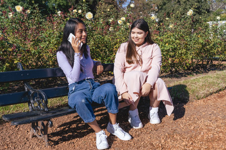 Two young women sitting on a park bench and one of them talking on her cell phone while the other girl watches her.の写真素材