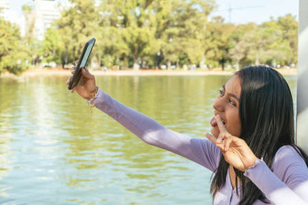 Attractive smiling Latina woman leaning on a white column in a public park taking a selfie