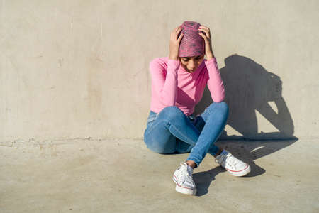Young woman with breast cancer sitting on the floor and leaning against a gray wall holding her head...