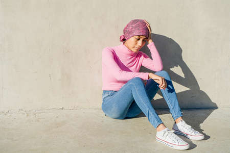 Young woman with breast cancer sitting on the floor leaning her head on her hand with a sad and...