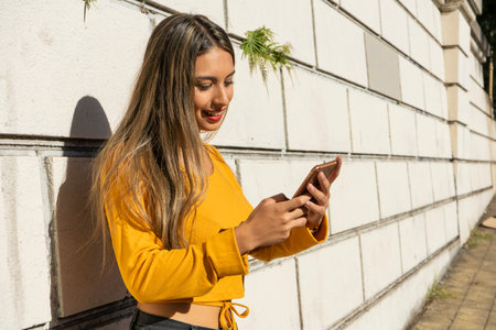 Portrait of a young Latina woman checking her smartphone or cell phone leaning against a wall....