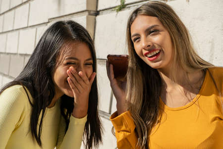 Two beautiful young Latina women giggling when listening to a funny audio message on their cell phone. Technology concept.の写真素材