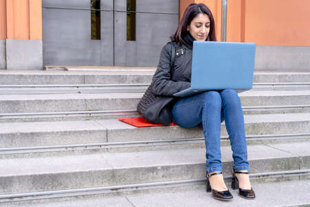 Portrait of a beautiful young Latina college woman working with her laptop on a staircase on a fall morning. Education, technology concept.の写真素材