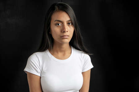 vertical medium close-up of a serious and beautiful black haired Latina woman wearing a white t-shirt on a black background with copy space.の写真素材